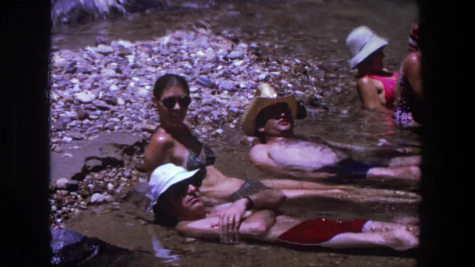 A small group in swimsuits, hats, and sunglasses rests in shallow Utah river water, framed by rocks and 8mm film grain.