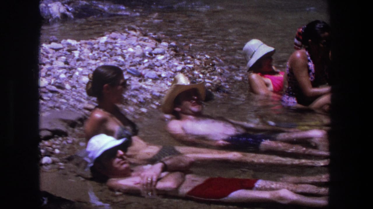 1969 Utah home-movie frame showing adults in swimsuits and sun hats relaxing in a shallow river with a rocky bank.