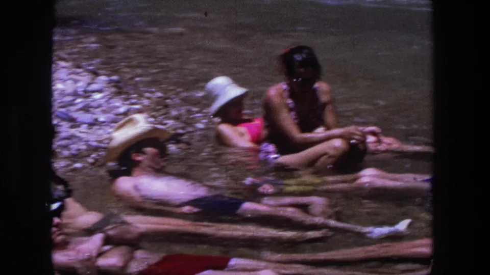Adults relax in a shallow Utah river in 1969, including reclining swimmers and a seated woman in patterned swimwear.