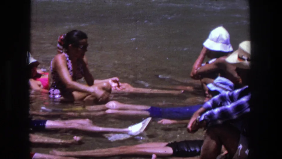 Home-movie frame from Utah in 1969 showing adults sitting and reclining in a shallow river under bright summer light.