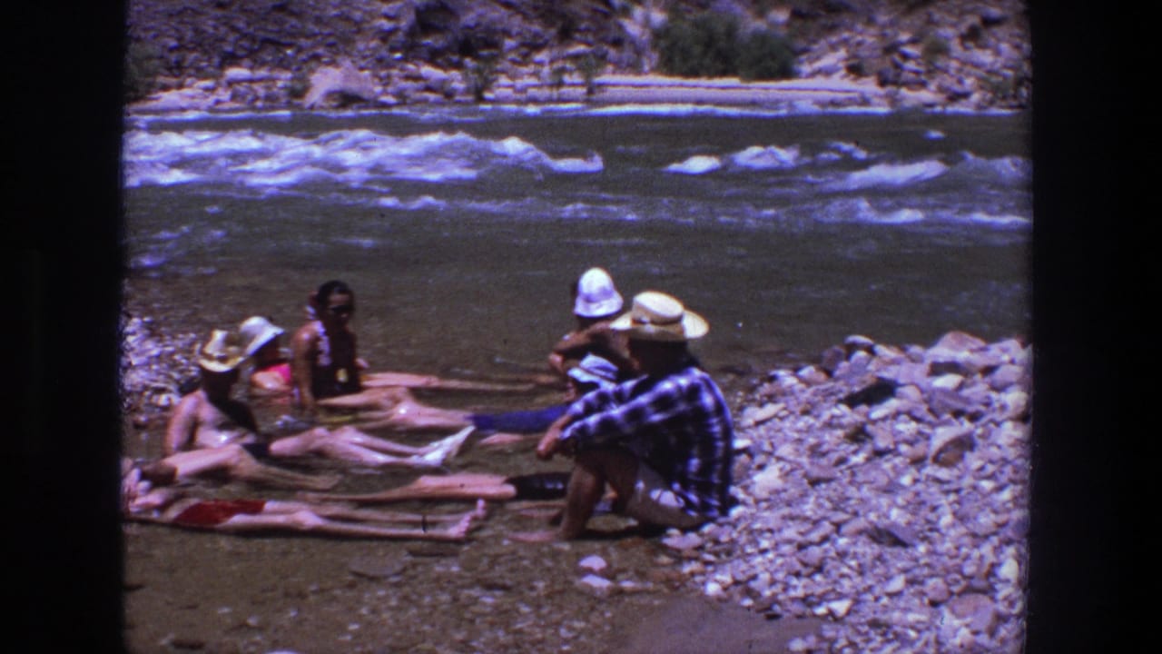 People sit and recline in shallow water beside a rocky riverbank.