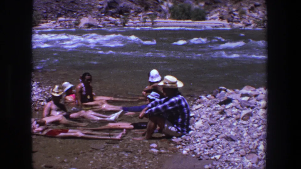 1969 Utah home-movie frame of adults relaxing in shallow river water near rocks and rushing water.