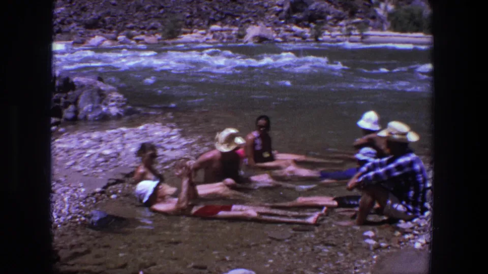 Adults relax in shallow water on a rocky Utah riverbank, filmed in 1969 on 8mm home-movie stock.