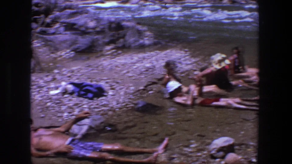 Wide 1969 Utah riverbank frame with adults lounging in shallow water and personal items on the rocky shore.