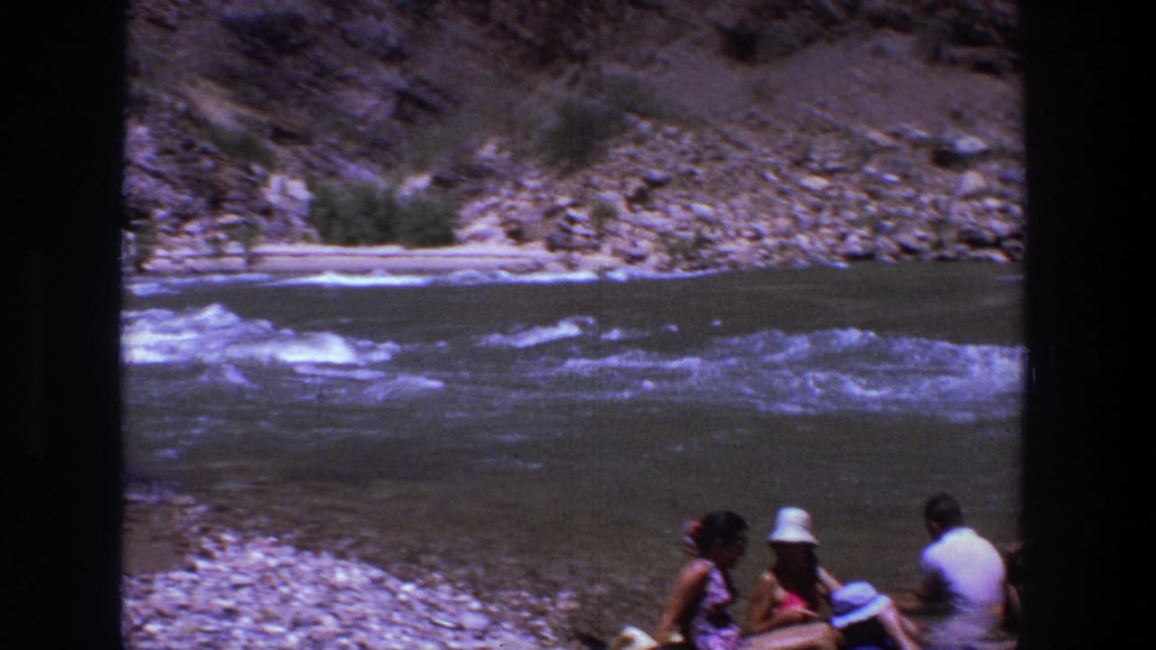 Small group sitting on a rocky riverbank beside rushing water and canyon walls.