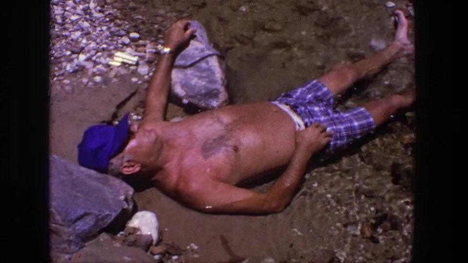 1969 Utah 8mm frame showing an adult man in plaid shorts and a blue cap reclining on stones and wet sand by shallow water.