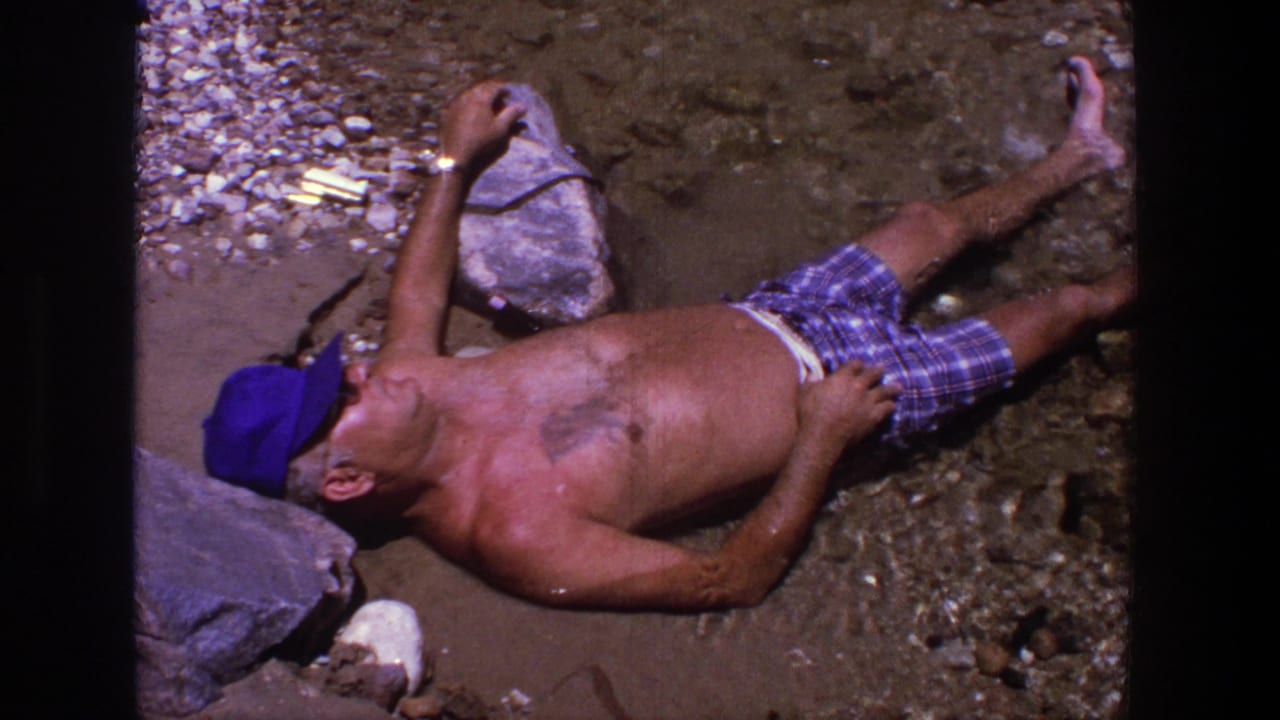 Shirtless man in blue cap lies on a rocky shore near shallow water.