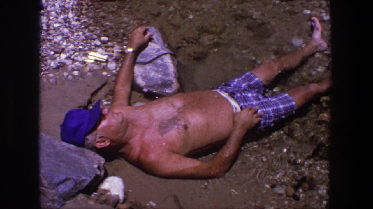 A man lies back on a rocky Utah shoreline, wearing a blue cap and plaid swim trunks in 1969 home-movie footage.