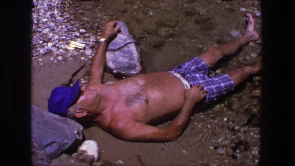 A man lies back on a rocky Utah shoreline, wearing a blue cap and plaid swim trunks in 1969 home-movie footage.