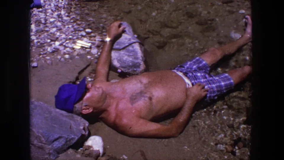 The 1969 Utah home-movie frame shows a shirtless man resting on a rocky beach with water and scattered stones around him.