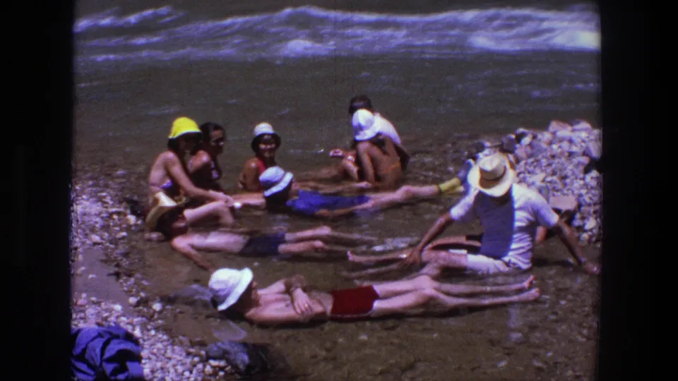 A small group sits and reclines in shallow shoreline water in supported 1969 Utah home-movie footage, with hats, swimwear, rocks, and waves visible.