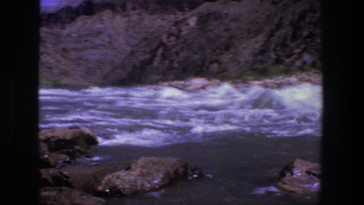 White water rushes past rocks below a rugged canyon wall.