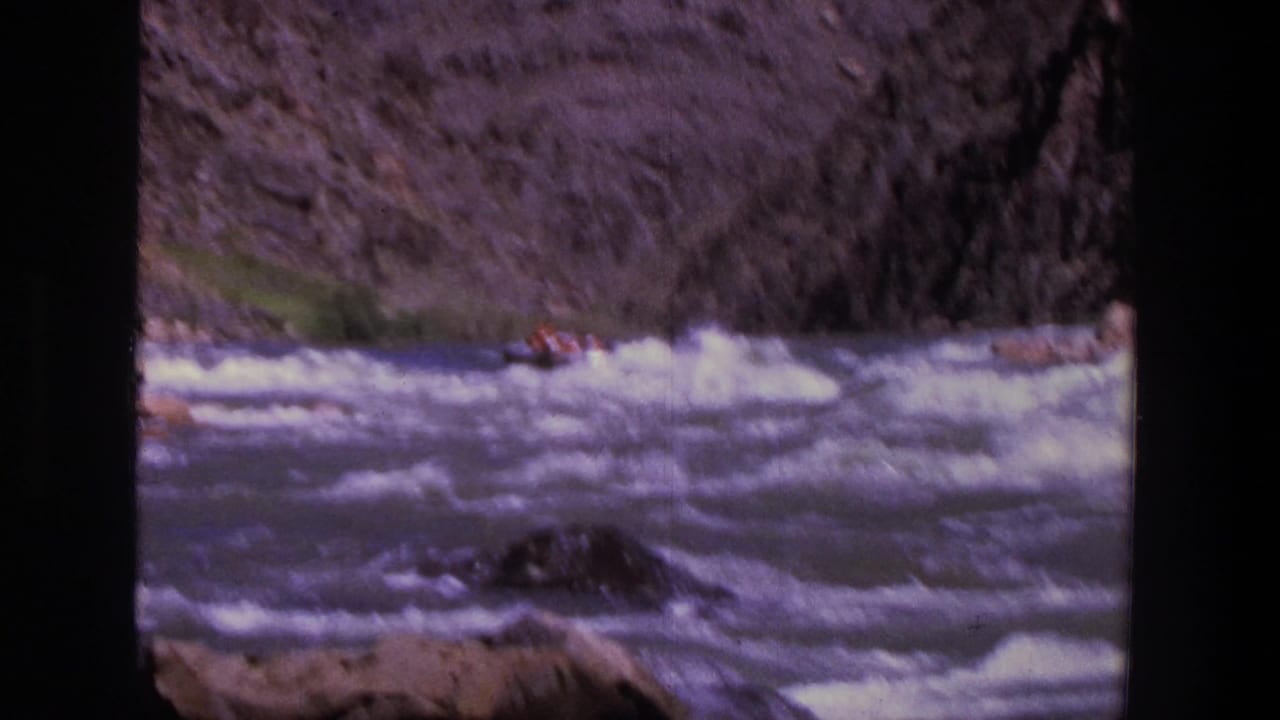 1969 Utah home-movie frame with a small passenger raft crossing bright rapids below steep canyon walls.