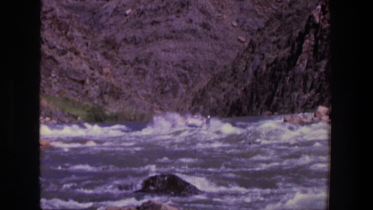 Foaming river rapids run through a rocky canyon with distant rafters near the center.