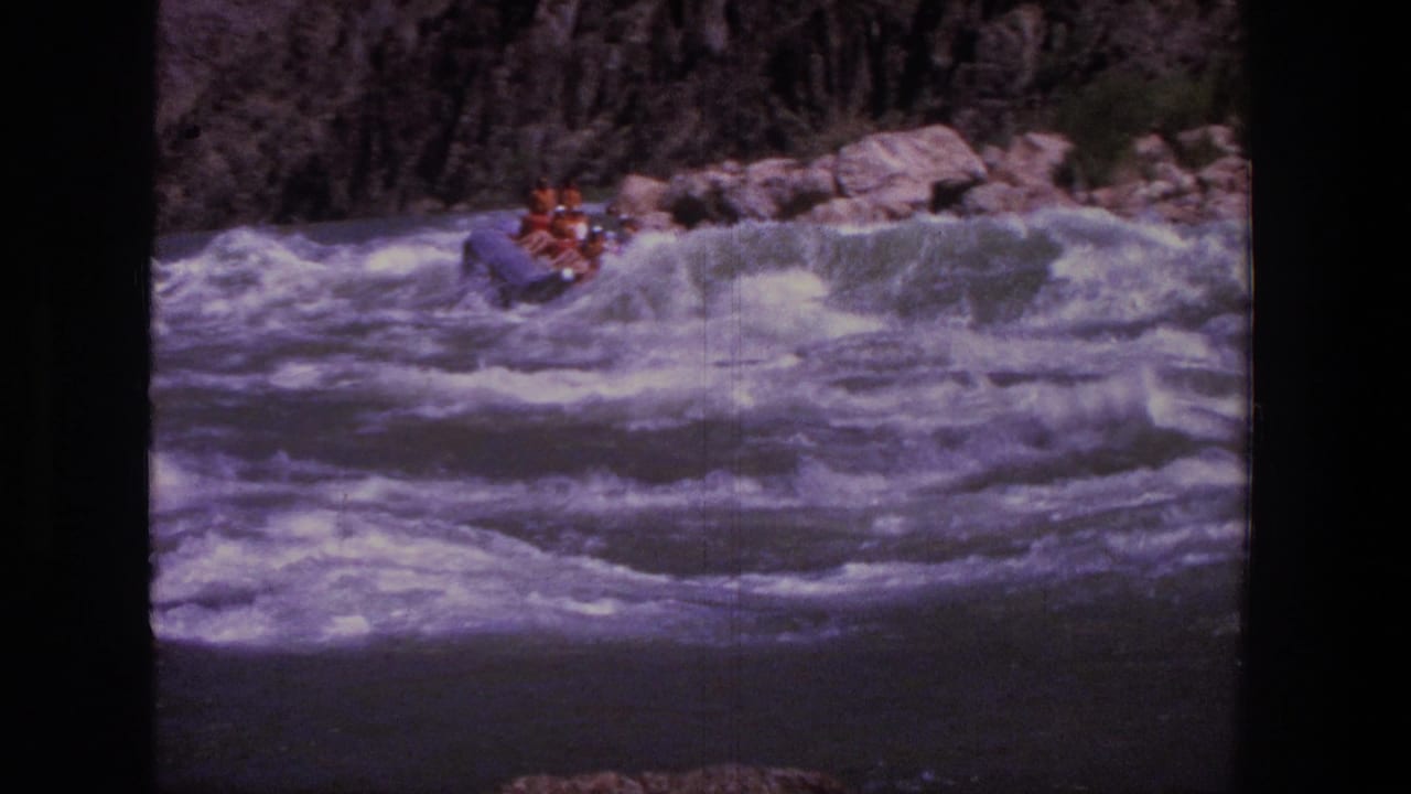 Inflatable raft with riders cresting foamy rapids near canyon rocks.
