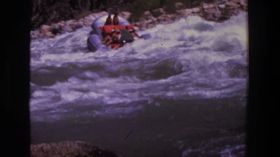 Close 1969 Utah frame of riders in a grey inflatable raft pushing through foamy rapids on a rocky river.
