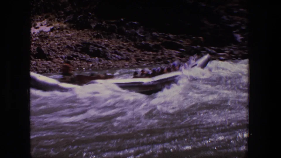 Inflatable raft moves through rough whitewater in Utah, with passengers indistinct and a rocky shoreline behind it.