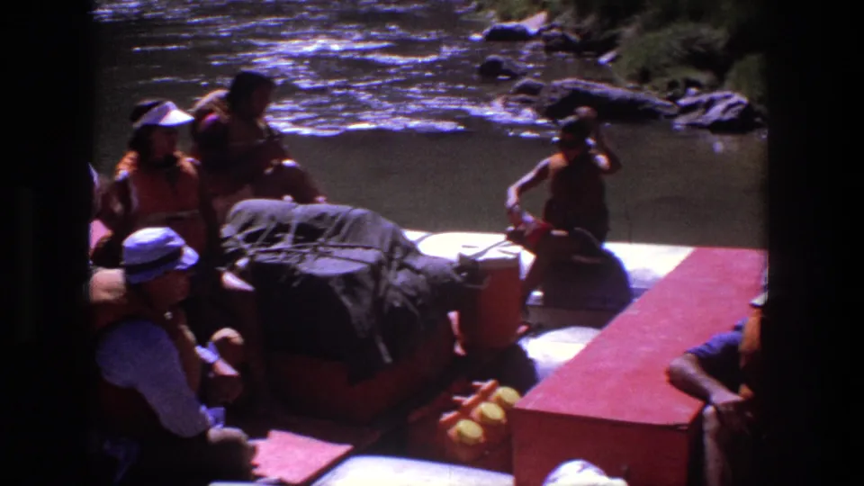 1969 Utah rafting scene with seated passengers, covered gear, and a standing person near the river shore.