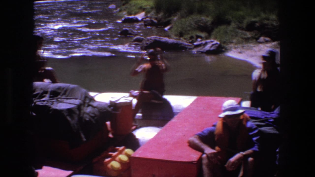 People in life vests sit on a red raft near a rocky riverbank.