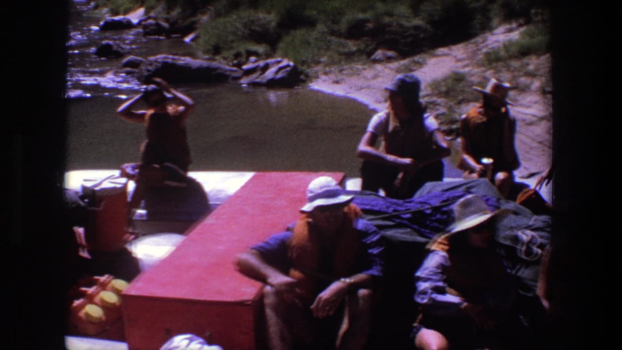 Several people sit on a red raft beside calm river water and rocks.