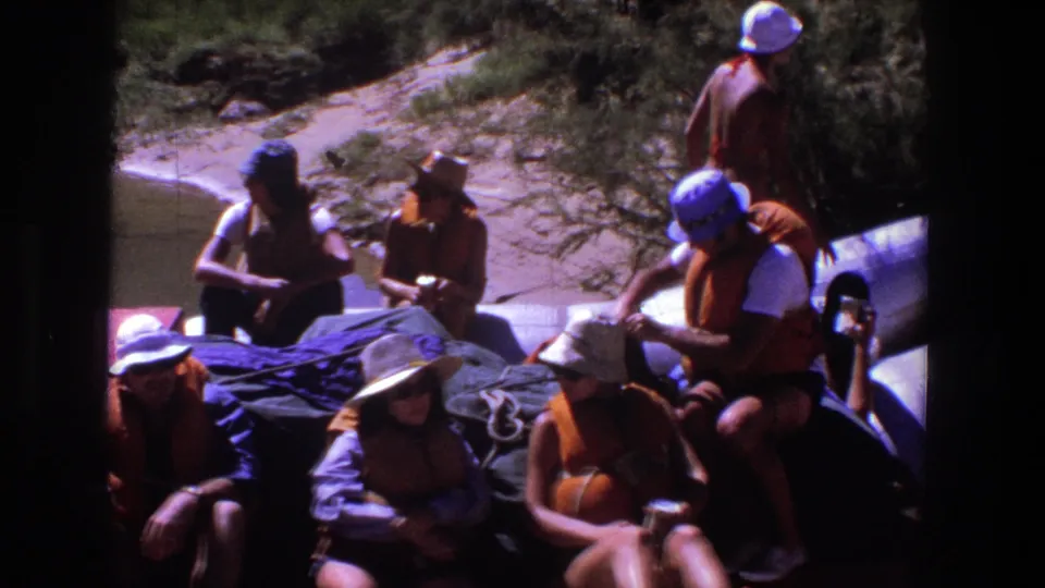 Adults on a red river raft in Utah, 1969, sit beside bundled gear and shallow water in bright sun.
