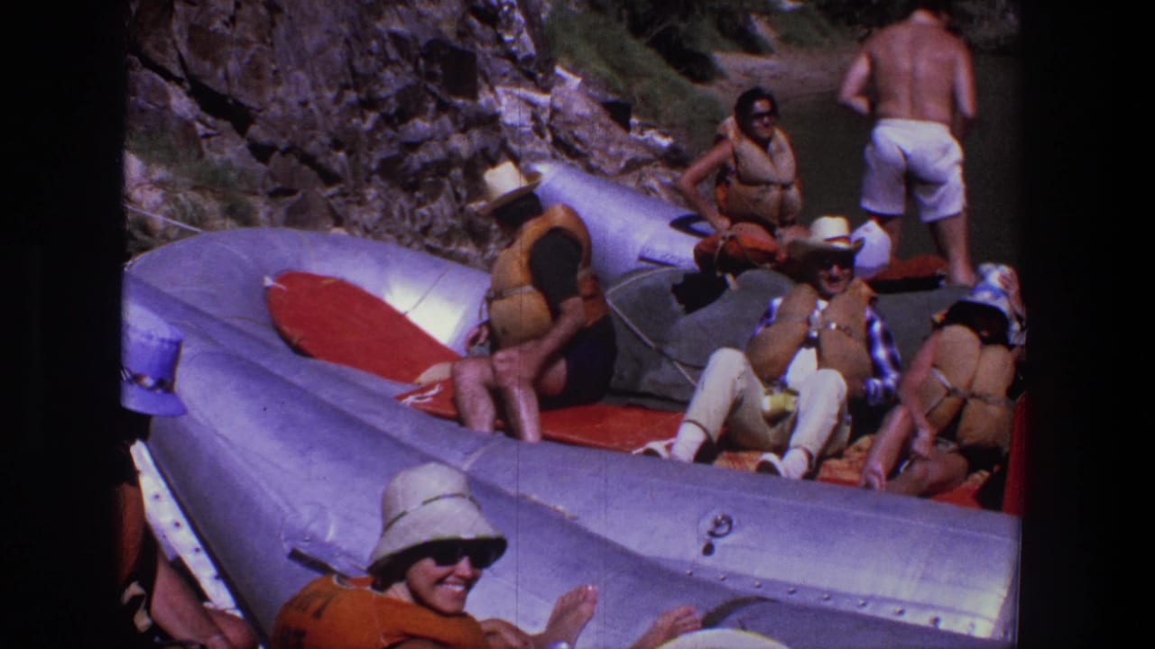 In supported 1969 Utah footage, adults in hats, sunglasses, and life vests rest in a silver inflatable raft near rocks and water.