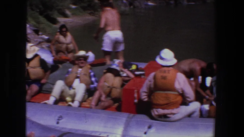 A home-movie frame shows adults resting in a silver inflatable raft, including a person in an orange life vest seen from behind.