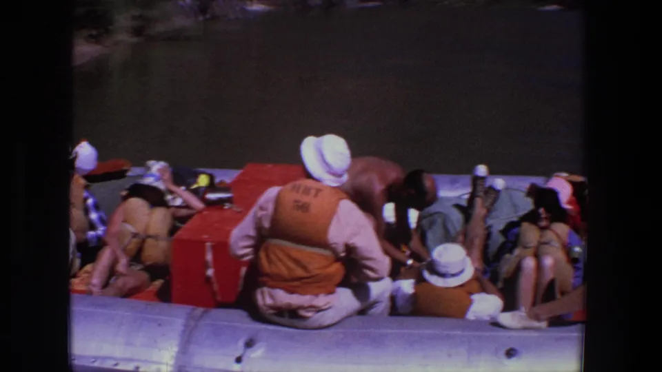 Adults in a silver inflatable raft rest near dark river water, with hats, orange life vests, and boating gear visible.