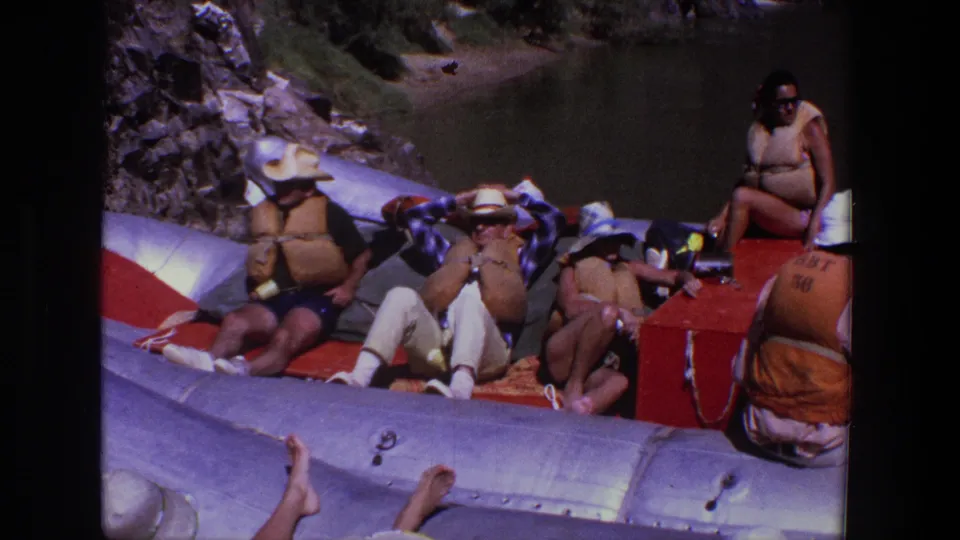 Utah river rafting scene with adults lounging on a large inflatable raft and a person seated near a red equipment box.
