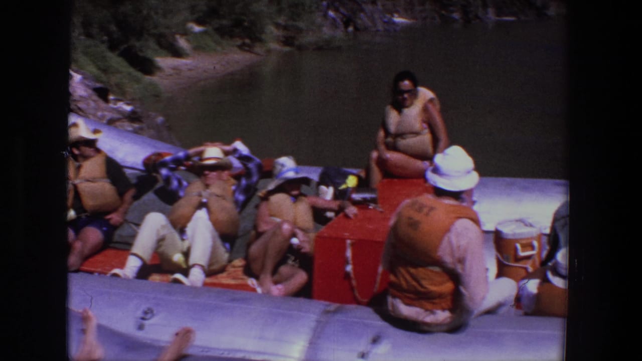 Adults in life vests lounge on an inflatable raft with coolers on a river.