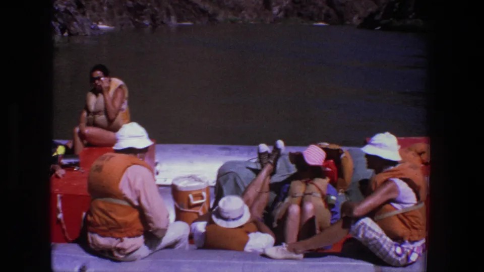 Home-movie frame from a 1969 Utah raft trip, showing several adults relaxing in orange life vests around coolers on calm river water.