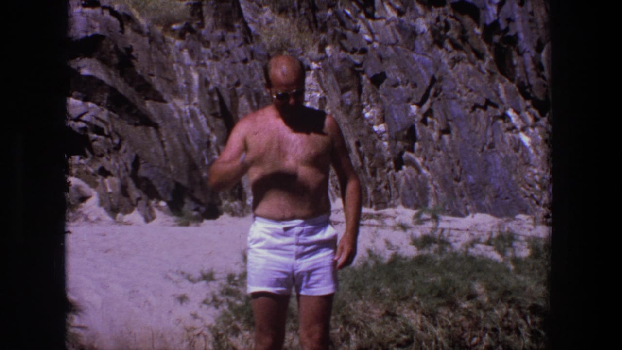 Shirtless adult man in white shorts stands on sandy ground before rock cliffs.