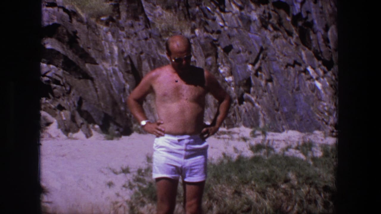 Shirtless man in white shorts stands with hands on hips before a rocky cliff.