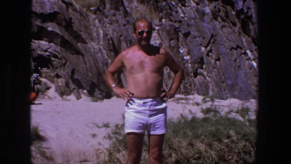 A shirtless adult man in sunglasses and white shorts faces the camera on a sandy Utah setting with rock cliffs behind him.