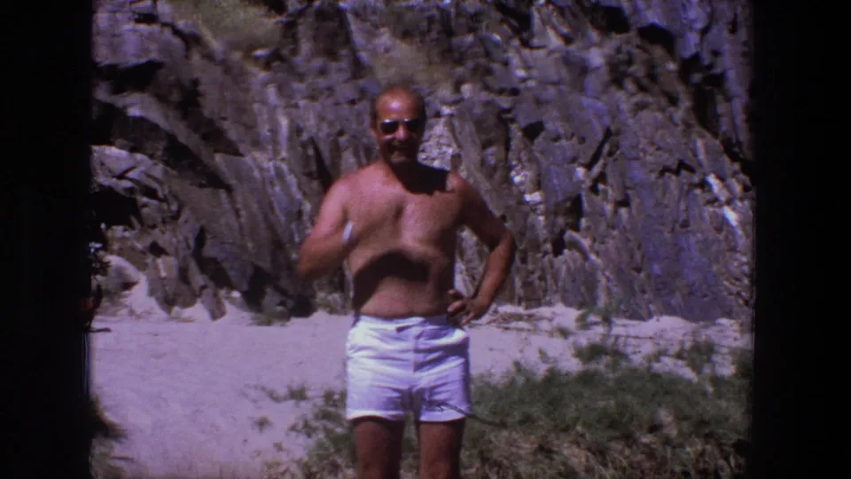 The man appears to adjust his sunglasses or gesture near his face while standing in white shorts before Utah rock cliffs.
