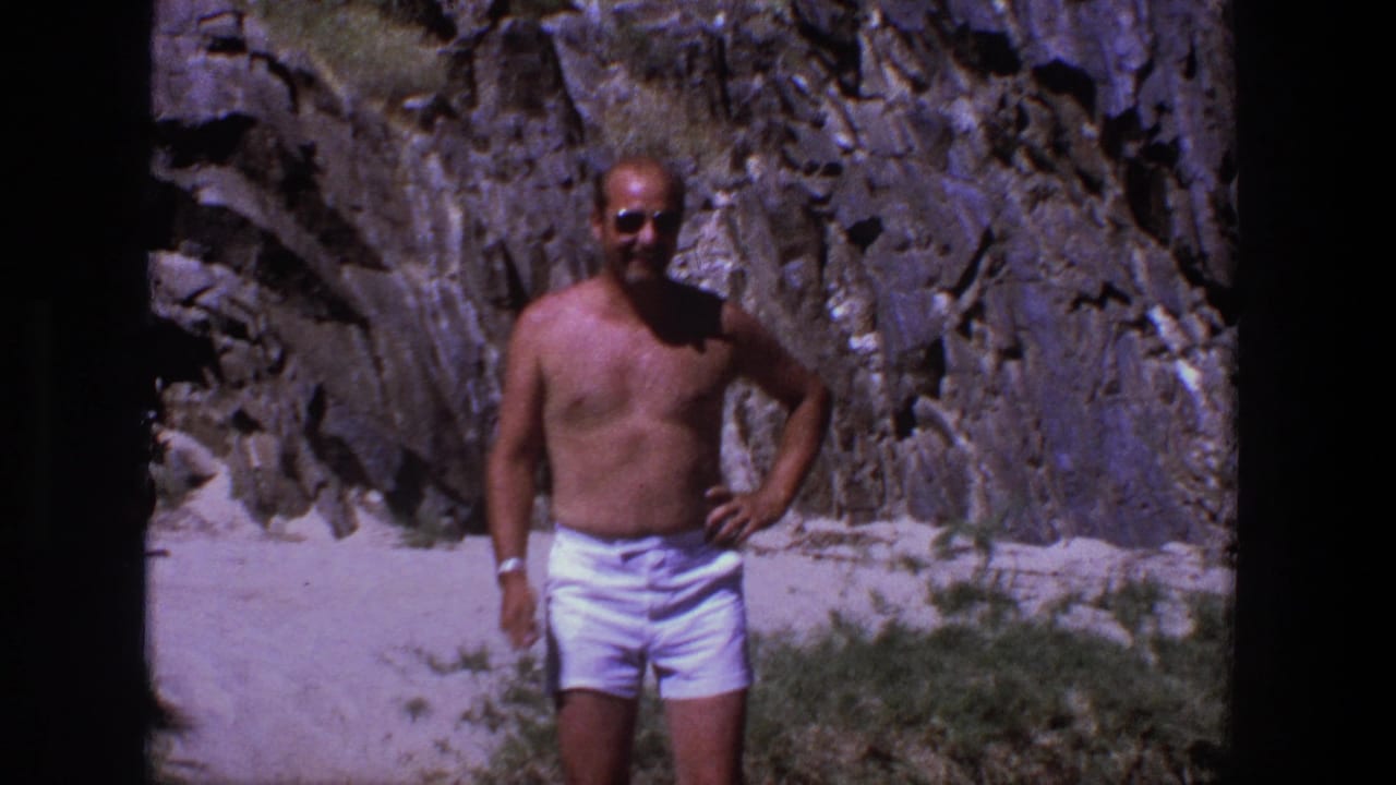 In 1969 Utah home-movie footage, a shirtless man in white shorts poses on a sunny sandy beach beneath rugged grey rock.