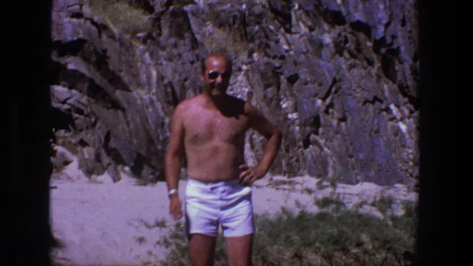 In 1969 Utah home-movie footage, a shirtless man in white shorts poses on a sunny sandy beach beneath rugged grey rock.