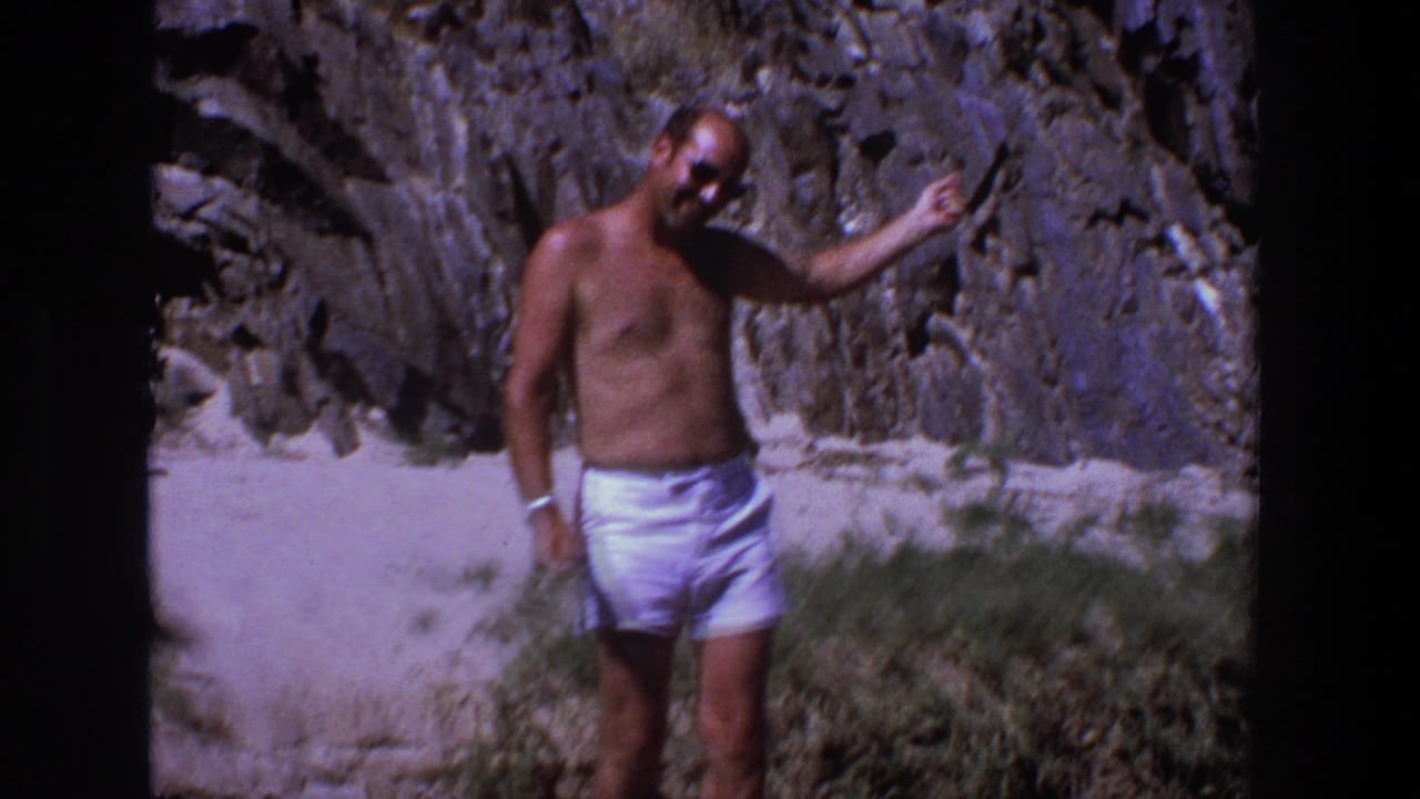 Man in white shorts gesturing beside a rugged rock wall on a sunny beach.