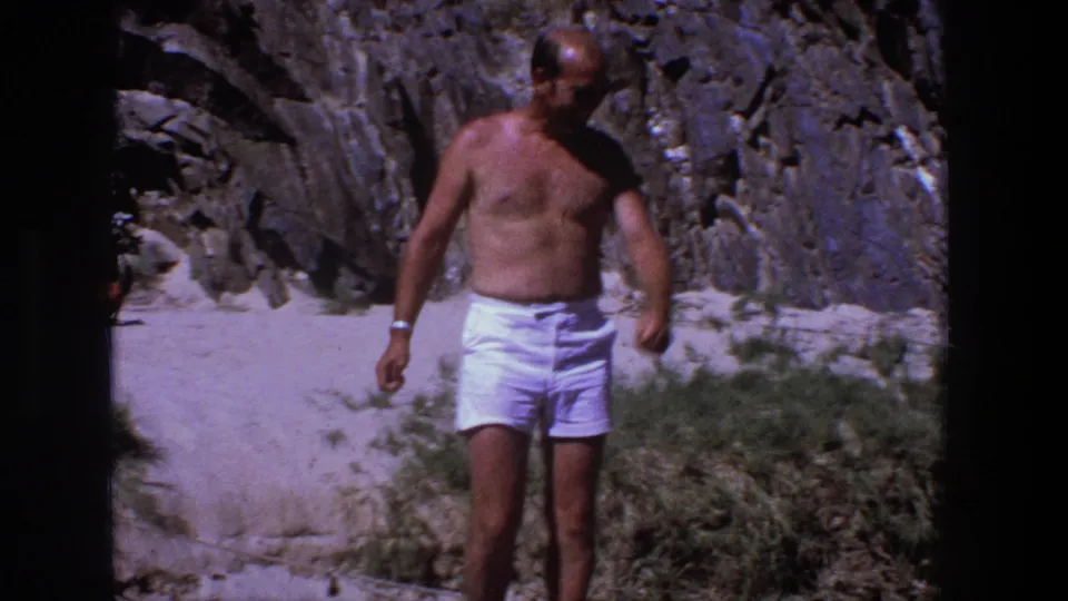 A shirtless man in white shorts walks near beach grass below a rugged grey rock face in the Utah 1969 clip.