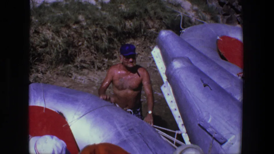 The man stands beside light-colored inflatable boats with red panels during a sunny Utah beach outing in 1969 footage.