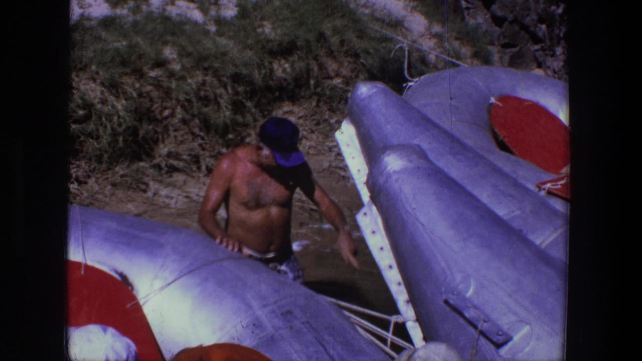 Shirtless man in a blue cap standing beside inflatable rafts on a riverbank.