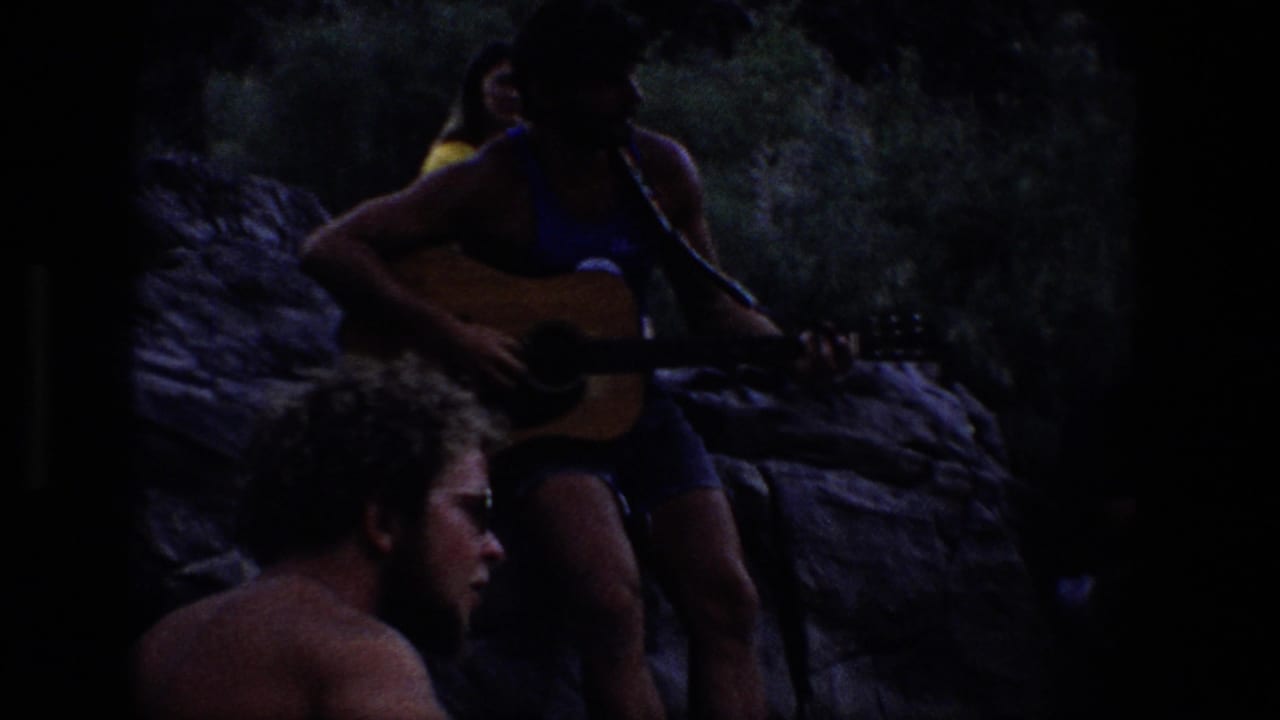 Dim 1969 Utah outdoor gathering with a man holding an acoustic guitar while companions sit nearby among rocks and trees.