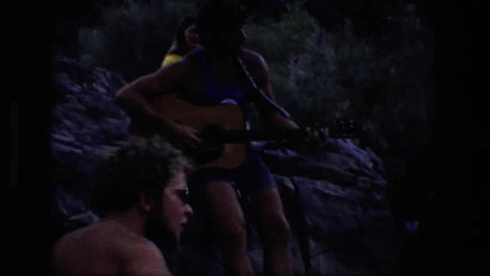 Dim 1969 Utah outdoor gathering with a man holding an acoustic guitar while companions sit nearby among rocks and trees.