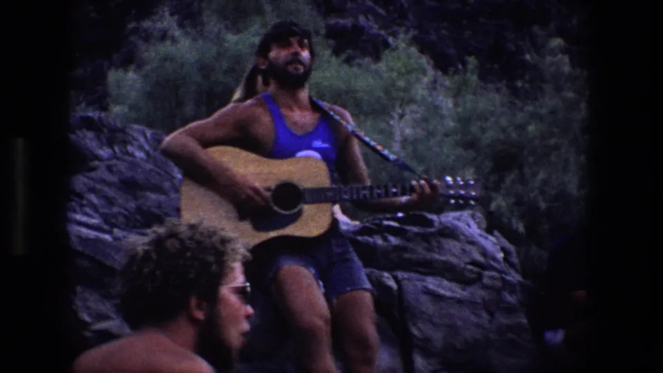 1969 Utah home-movie frame showing a man in a blue tank top playing acoustic guitar beside a seated friend outdoors.