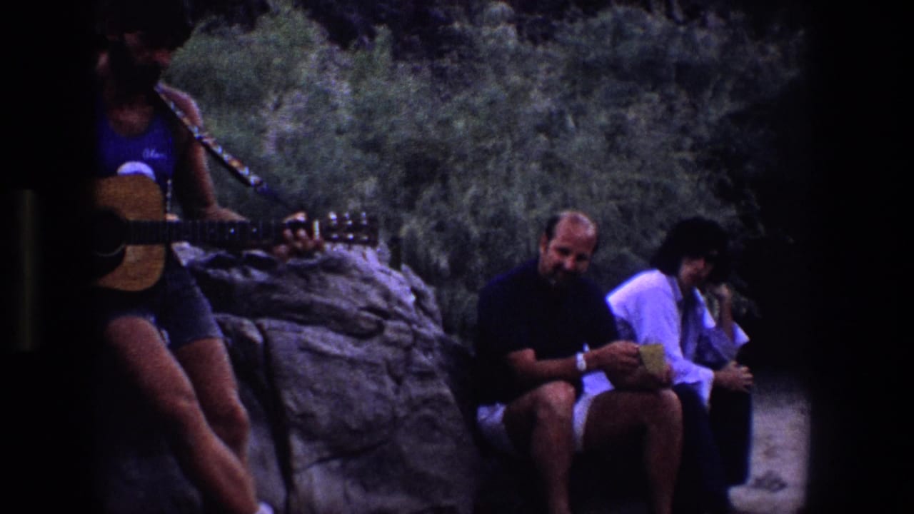 Friends sit on rocks outdoors while a man at left plays acoustic guitar.