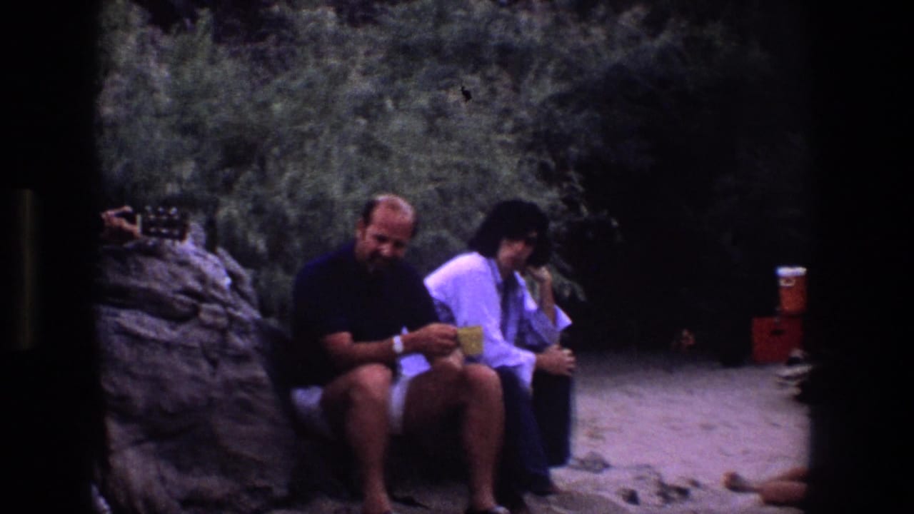 Two seated friends hold cups near a rock ledge and wooded background.