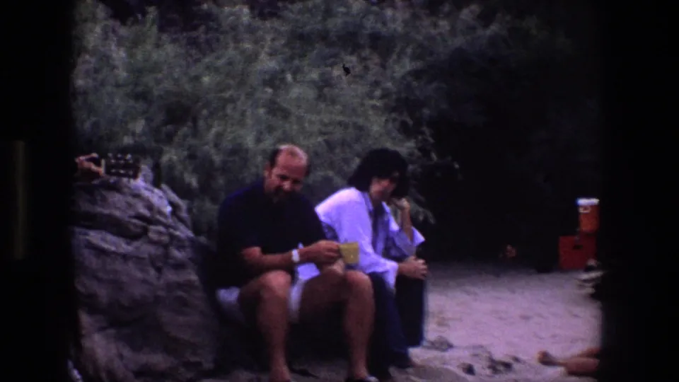 1969 Utah 8mm frame showing two adults seated outdoors with cups, while an acoustic guitar neck enters from the left edge.