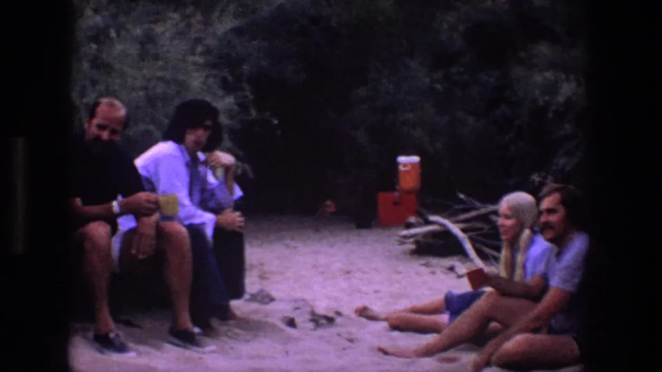 1969 Utah 8mm frame of friends seated on rocks and sandy ground, with a red cooler in the background and relaxed conversation around the music circle.