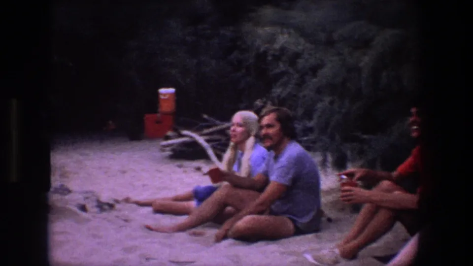 1969 Utah 8mm frame showing seated friends on sandy ground beside brush, logs, and a red drink cooler during an informal outdoor gathering.