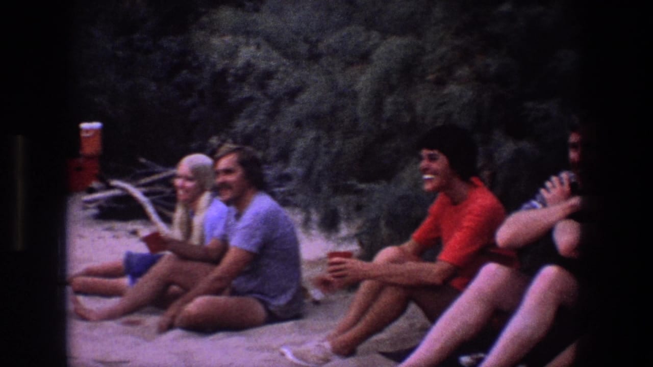 Young adults sit on sand near brush, laughing and holding cups.